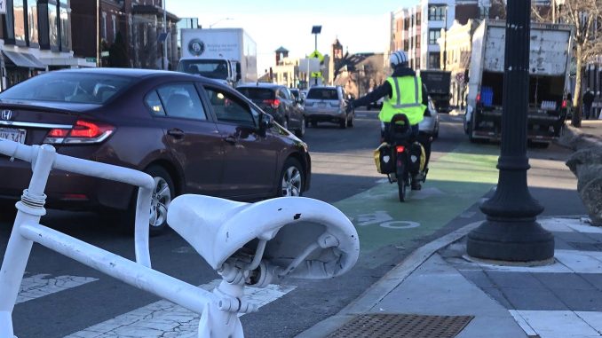A photo of the Porter Square intersection. In the foreground is a white ghost/memorial bike. In the background, a cyclist wearing a high visibility fluorescent vest is riding in the bike lane, and up ahead a truck is parked in the bike lane,